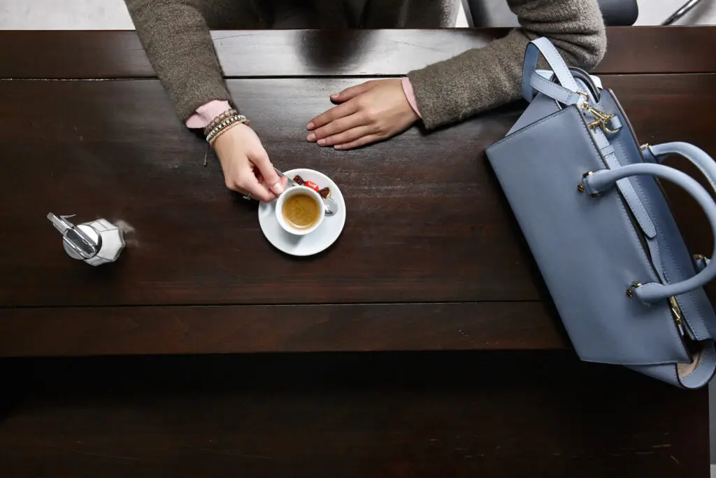 Eine Hand greift nach einer Tasse Espresso auf einem weißen Unterteller, während eine elegante Handtasche und ein Zuckergefäß auf einem dunklen Holztisch platziert sind.