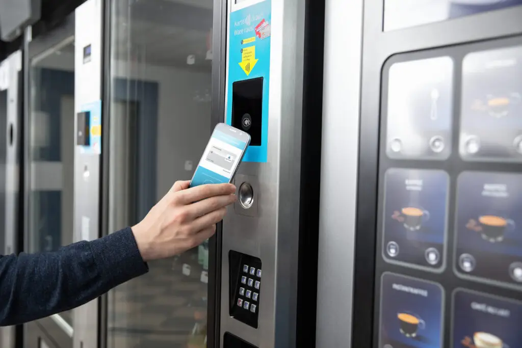 Person scannt ihr Smartphone an einem modernen Verkaufsautomaten zur Bezahlung, der eine digitale Benutzeroberfläche zeigt. Der Automat bietet verschiedene Snacks und Getränke an.