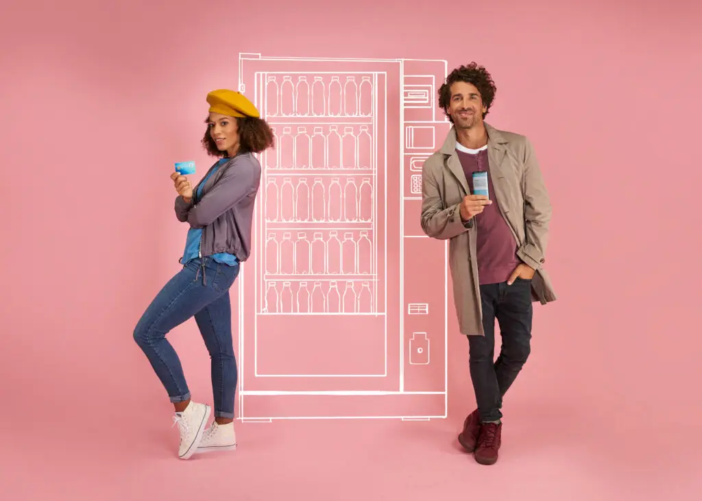 Junge Frau und junger Mann stehen vor einem rosa Hintergrund, lächelnd und jeweils mit einem Smartphone in der Hand. Im Hintergrund ist eine stilisierte Skizze eines Getränkeautomaten zu sehen. Die Frau trägt eine gelbe Mütze und eine lockere Jacke, während der Mann in einem beigen Mantel gekleidet ist.