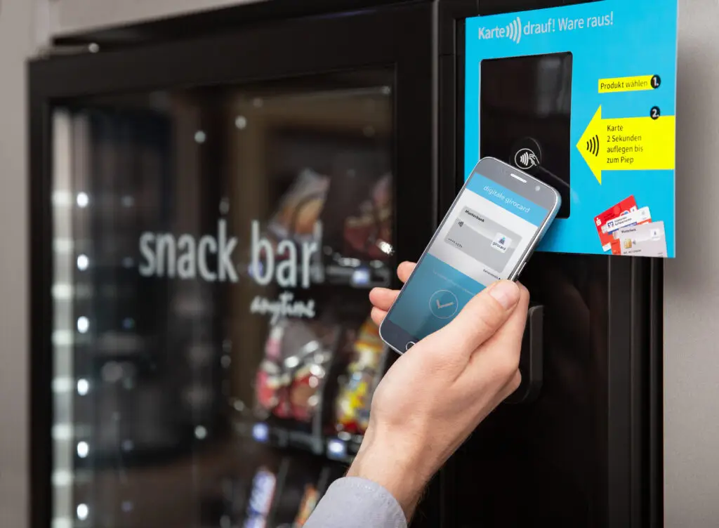 Hand, die ein Smartphone mit digitaler Girocard an einen modernen Snackautomaten hält, während das Display die Zahlungsbestätigung anzeigt. Im Hintergrund sind verschiedene Snacks sichtbar, und eine Anleitung zur Nutzung des Automaten ist gut lesbar.