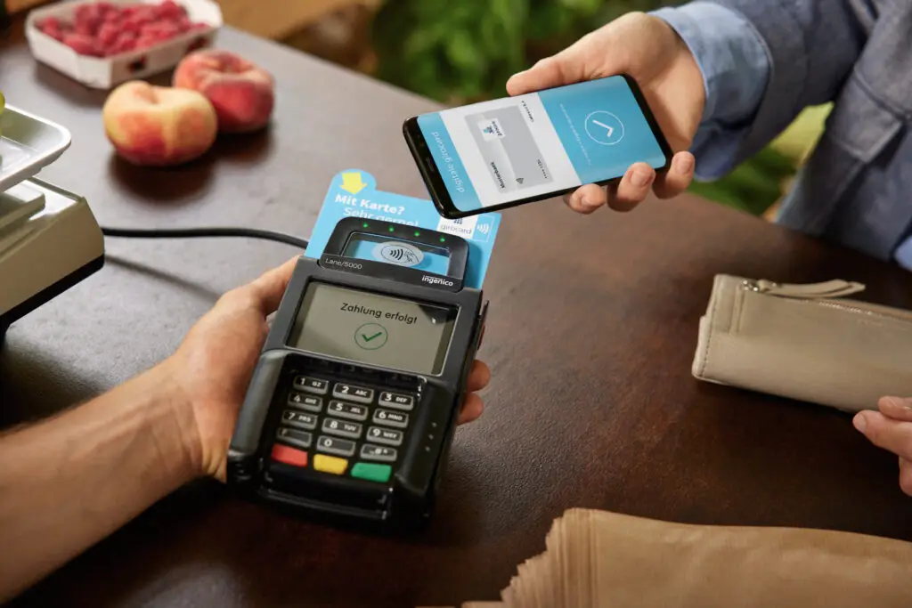 Zahlung per Smartphone an einem kontaktlosen Kartenlesegerät, das den erfolgreichen Abschluss der Transaktion anzeigt, während frisches Obst im Hintergrund sichtbar ist.