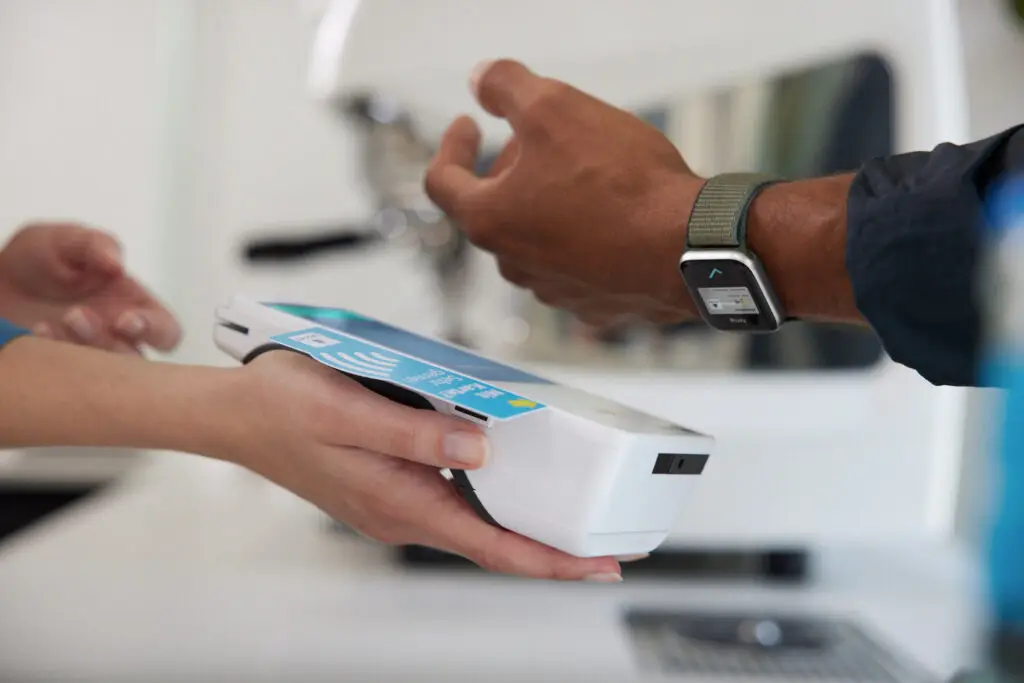 Zahlungsvorgang mit kontaktlosem Bezahlsystem, bei dem eine Person ein mobiles Zahlungsterminal in der Hand hält, während eine andere Person ihr Handgelenk mit einer Smartwatch zur Zahlung näherbringt.