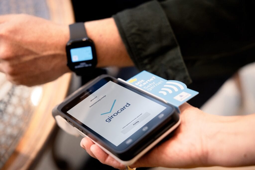 Person führt eine kontaktlose Zahlung mit einer girocard über ein mobiles Zahlungsterminal durch, während eine Smartwatch am Handgelenk sichtbar ist.