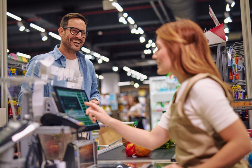 Lächelnder Kunde an der Kasse eines Supermarktes, während die Kassiererin ihn bedient und auf den Bildschirm zeigt. Im Hintergrund sind Regale mit verschiedenen Produkten zu sehen.